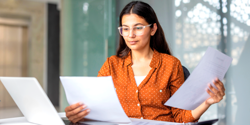 Mulher de óculos, sentada em uma cadeira com folhas de papel nas mãos.