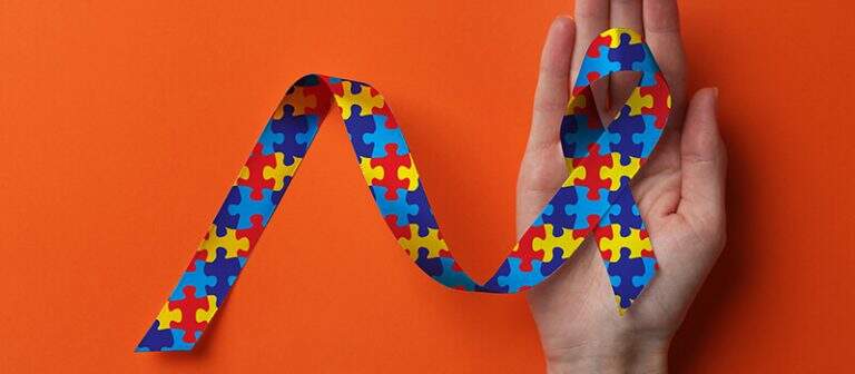 Mão segurando a fita da conscientização sobre o autismo, estampada com peças de quebra-cabeça amarelas, vermelhas e azuis, sobre um fundo laranja radiante.
