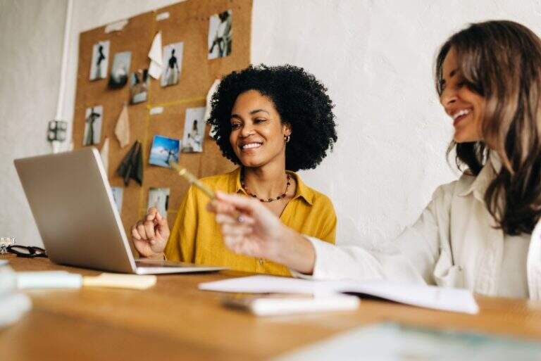 Duas mulheres sorridentes trabalhando juntas em um ateliê criativo. Uma mulher negra de camisa amarela olha para a tela de um notebook enquanto sua colega aponta algo com uma caneta.
