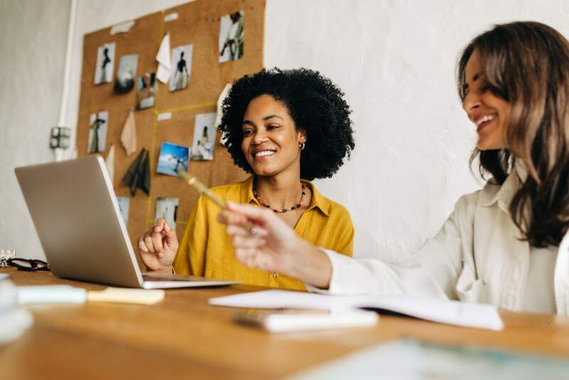 Duas mulheres sorridentes trabalhando juntas em um ateliê criativo. Uma mulher negra de camisa amarela olha para a tela de um notebook enquanto sua colega aponta algo com uma caneta.