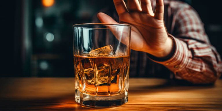 copo de bebida alcoólica em uma mesa e uma pessoa, apenas a mão dela aparente, recusando o copo de bebida.